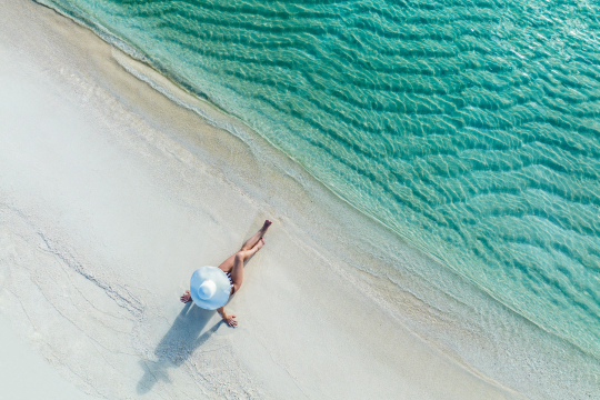 Frau mit weißem Sonnenhut sitzt an einem weißem Sandstrand mit türkisfarbenen Wasser